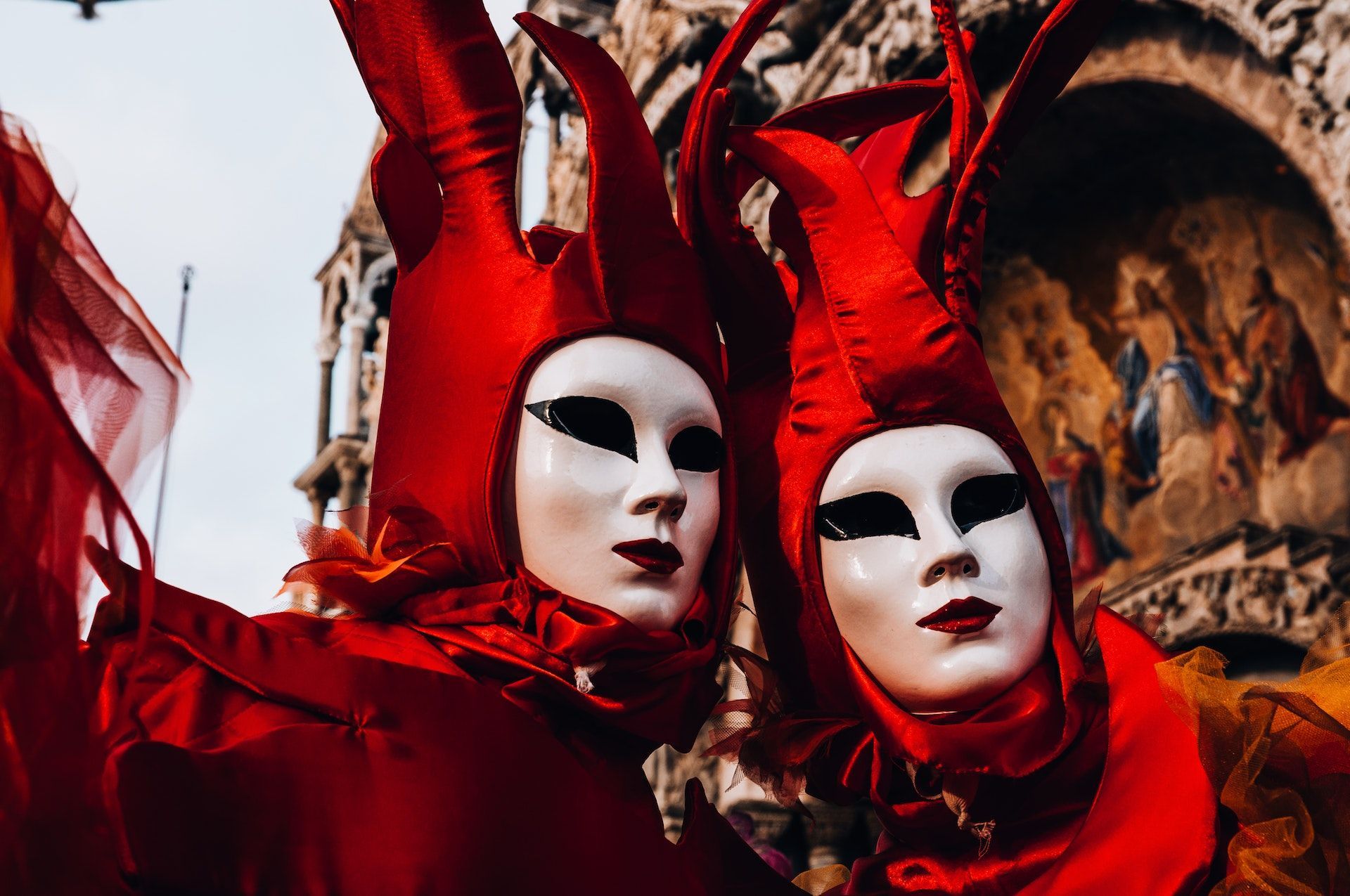 A pair of figures at a carnival, wearing elaborate red costumes and unsettling, almost featureless white masks
