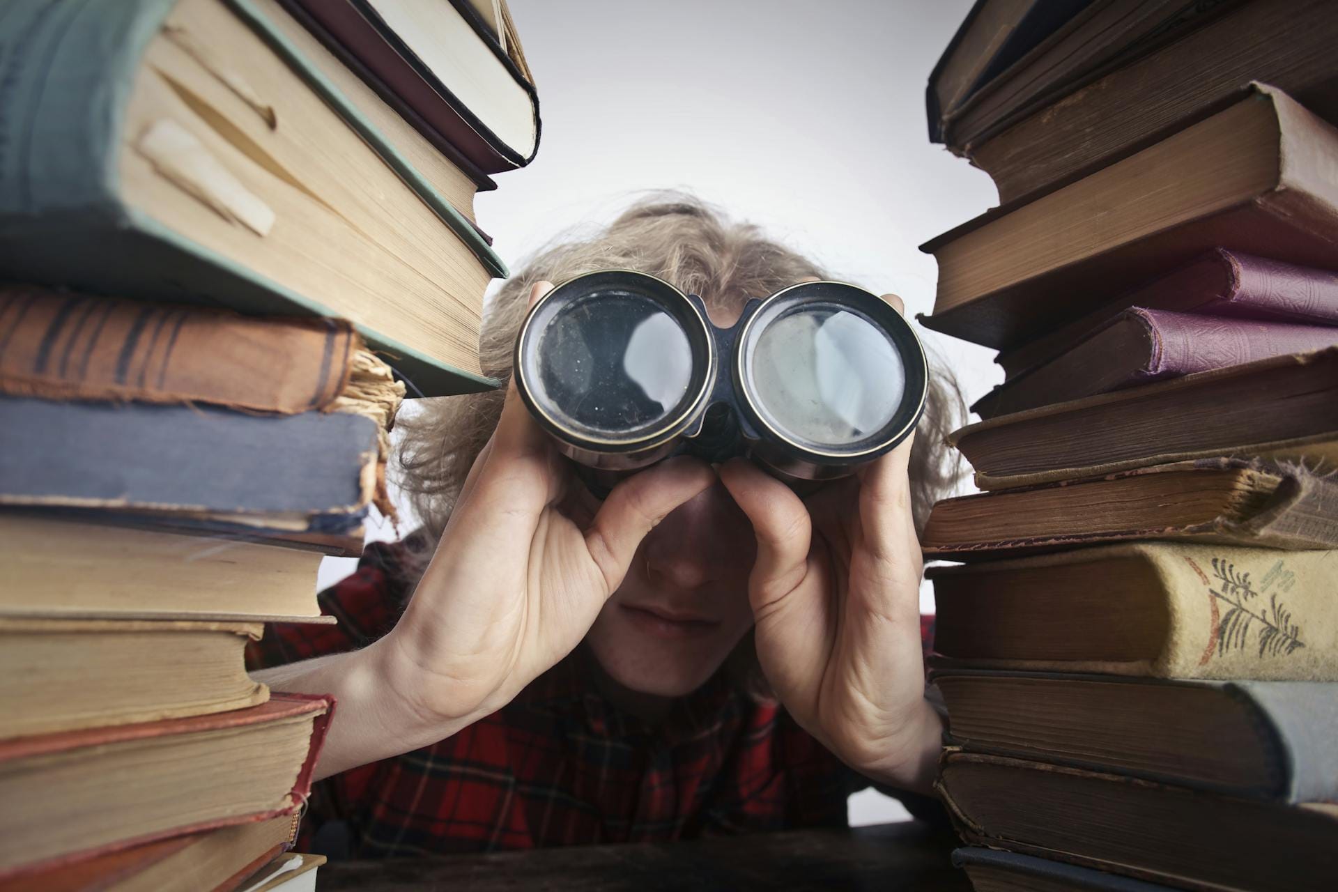 A man searching through binoculars, surrounded by books