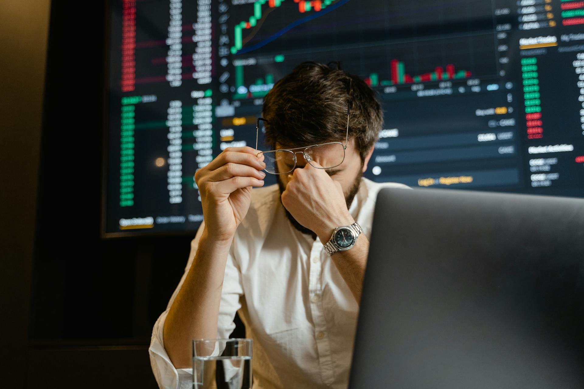 A man pinches the bridge of his nose, while surrounded by computer screens showing financial charts