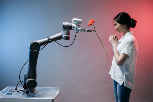 An industrial robot arm presents a carnation to a woman, who seems pleased by this.